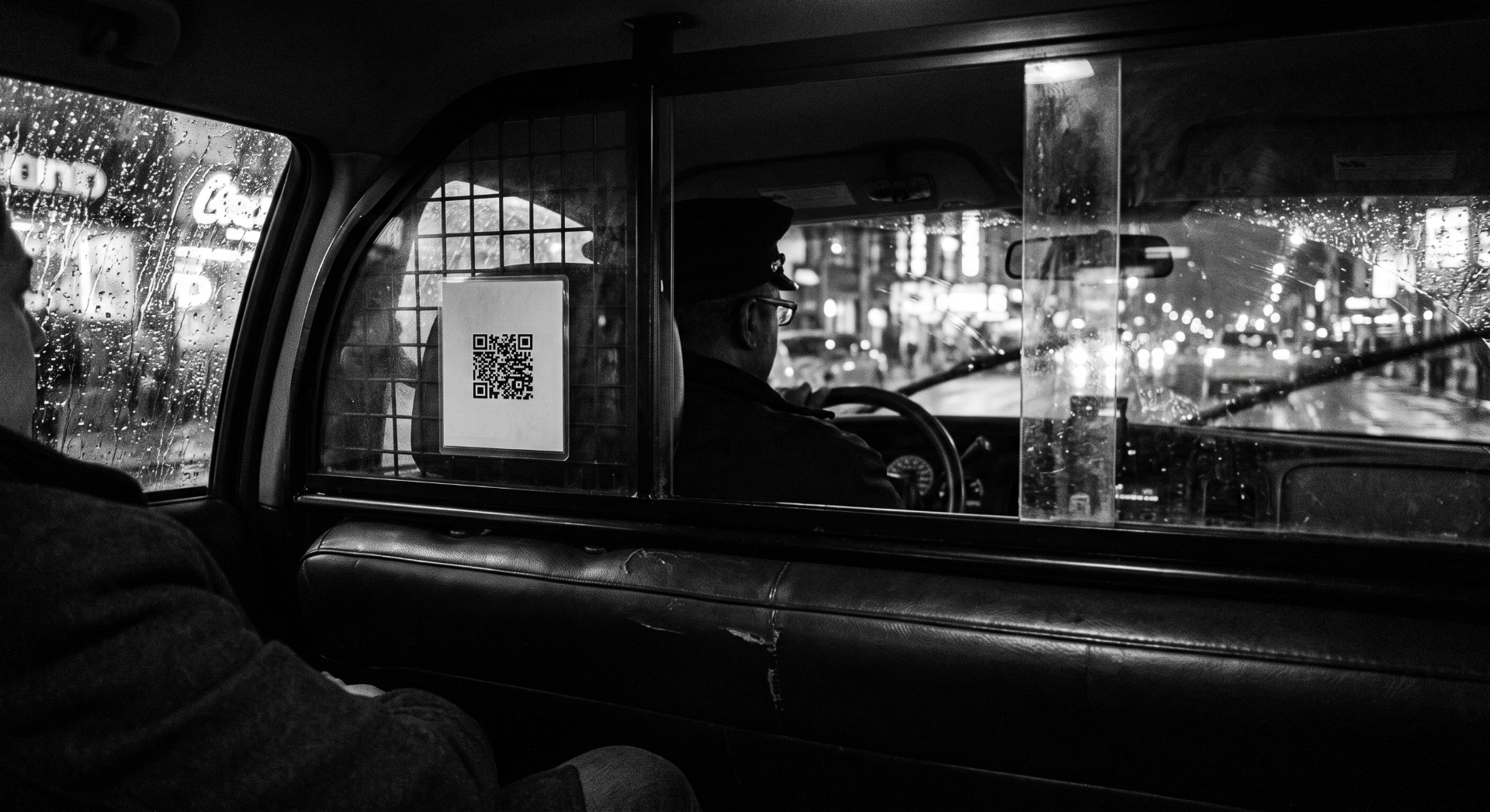 A QR code taped to the partition of a taxi cab at night, seen from the back seat